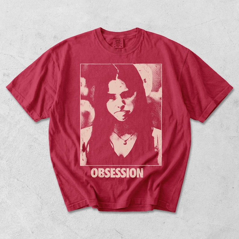 May include: A red short-sleeve t-shirt featuring a rectangular graphic print of a person with long hair wearing a necklace. The print is in a lighter shade of red and includes the word OBSESSION printed in capital letters below the image.