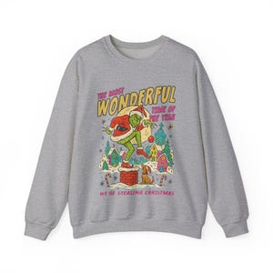 May include: A gray sweatshirt featuring a graphic of a green character in a Santa hat, standing on a chimney. The text reads "THE MOST WONDERFUL TIME OF THE YEAR" and "WE'RE STEALING CHRISTMAS". The design includes a snowy village scene.