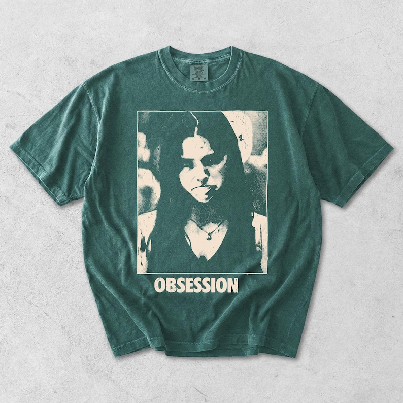 May include: A dark green short-sleeve t-shirt featuring a cream-colored graphic print of a person with long hair and a necklace. The word OBSESSION is printed in a distressed, bold, sans-serif font directly below the central portrait graphic.