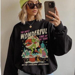 May include: Black sweatshirt with a graphic of the Grinch stealing Christmas. The text reads "THE MOST WONDERFUL TIME OF THE YEAR" and "WE'RE STEALING CHRISTMAS". The Grinch is depicted in a Santa suit with a sack of gifts.