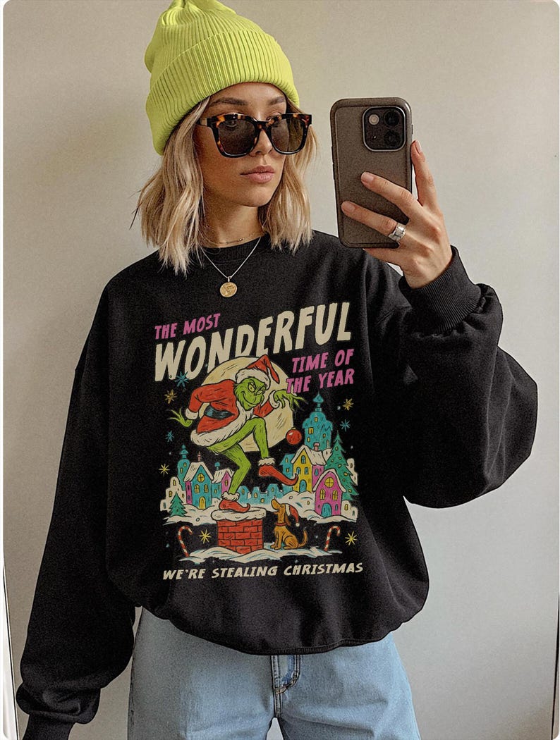 May include: Black sweatshirt with a graphic of the Grinch stealing Christmas. The text reads "THE MOST WONDERFUL TIME OF THE YEAR" and "WE'RE STEALING CHRISTMAS". The Grinch is depicted in a Santa suit with a sack of gifts.