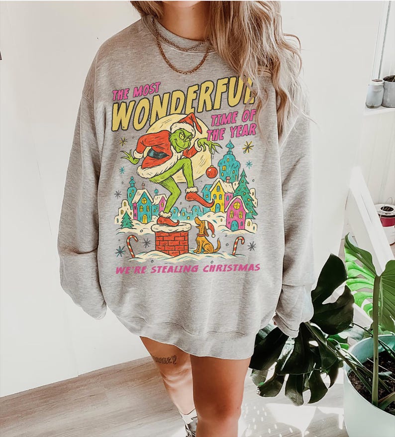 May include: A gray sweatshirt with a colorful graphic featuring a green character in a Santa hat, with the text "THE MOST WONDERFUL TIME OF THE YEAR" and "WE'RE STEALING CHRISTMAS". The design includes houses, trees, and a dog.
