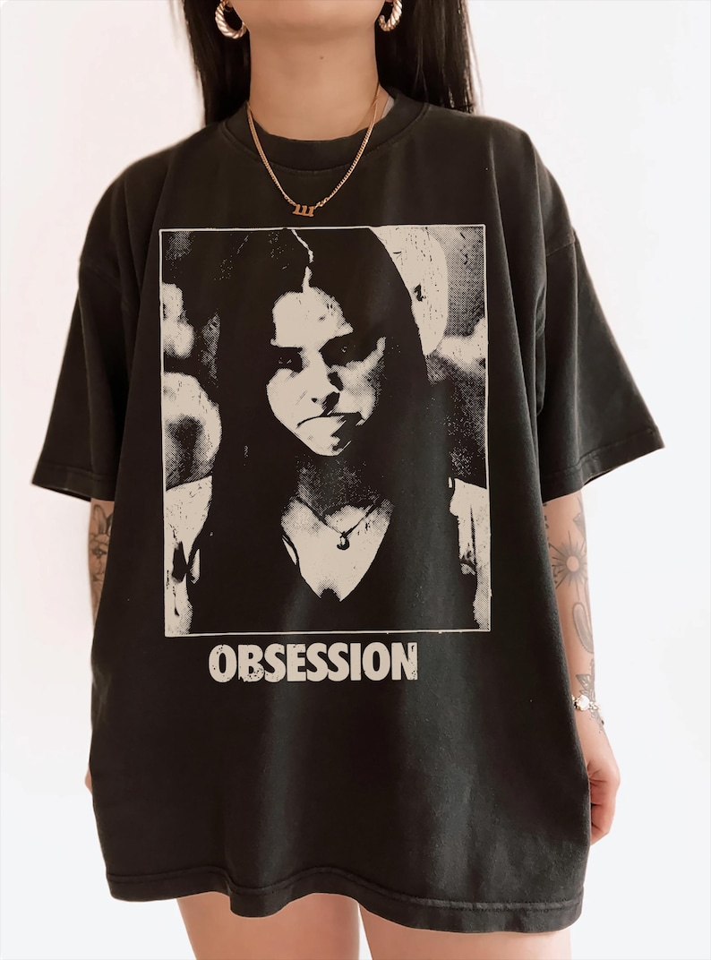 May include: A black short-sleeve t-shirt featuring a rectangular graphic print of a person with long dark hair. The print is in a high-contrast black and white style. Below the image, the word OBSESSION is printed in bold white capital letters.