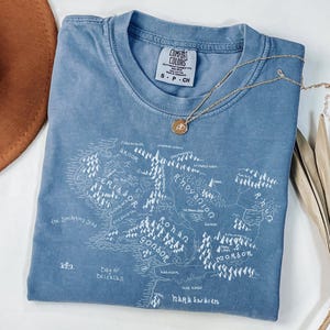 May include: A blue Comfort Colors t-shirt with a white map graphic and the text "Rohan" and "Gondor". A gold necklace with a pendant is draped across the shirt. The shirt is folded on a white surface.