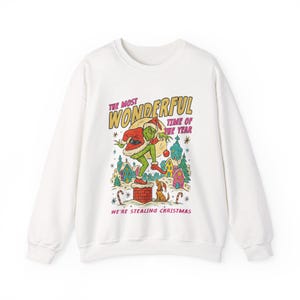 May include: White sweatshirt with a colorful graphic of a green character in a Santa suit, standing on a chimney. The text reads "THE MOST WONDERFUL TIME OF THE YEAR" and "WE'RE STEALING CHRISTMAS".
