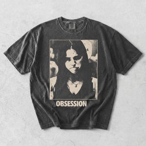 May include: A charcoal grey short-sleeve t-shirt with a vintage-style wash. The center features a rectangular graphic print of a person with long dark hair wearing a necklace, with the word OBSESSION printed in bold white capital letters below.