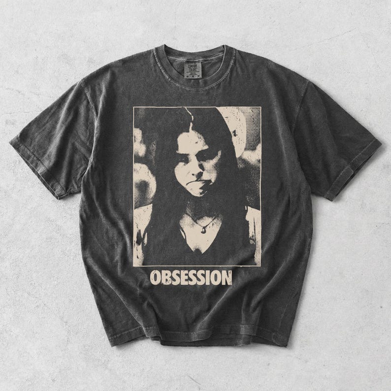 May include: A charcoal grey short-sleeve t-shirt with a vintage-style wash. The center features a rectangular graphic print of a person with long dark hair wearing a necklace, with the word OBSESSION printed in bold white capital letters below.