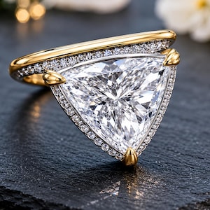 Pode incluir: Um anel de ouro com uma grande pedra central transparente em lapidação trillion. A cravação é adornada com um halo de pequenas pedras em pavé ao longo da estrutura triangular e do aro, fixada por três garras de ouro nos cantos.