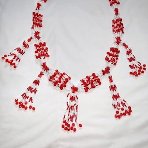 Op de afbeelding: Een ketting van witte en rode kralen. Het ontwerp bestaat uit clusters van rode kralen afgewisseld met witte kralen. De ketting heeft verschillende kralenkwasten. De ketting wordt getoond tegen een witte achtergrond.
