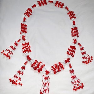 Op de afbeelding: Een ketting van witte en rode kralen. De ketting heeft een centraal ontwerp met meerdere kralenkwasten. De kralen zijn klein en rond, en de kwasten zijn lang en vloeiend, wat een elegante uitstraling cre&euml;ert.