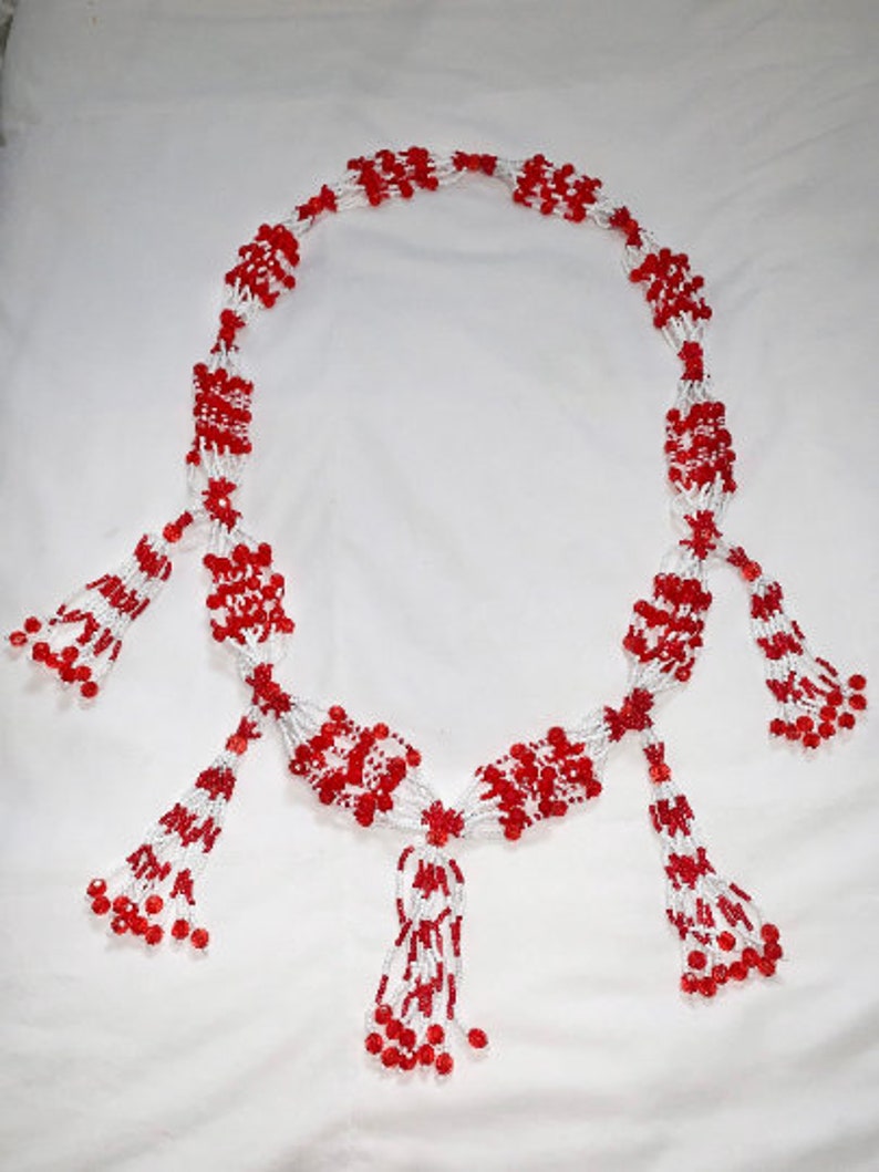 Op de afbeelding: Een ketting van witte en rode kralen. De ketting heeft een centraal ontwerp met meerdere kralenkwasten. De kralen zijn klein en rond, en de kwasten zijn lang en vloeiend, wat een elegante uitstraling cre&euml;ert.