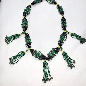 May include: A necklace made of black, green, and silver beads. The design includes multiple beaded sections linked by yellow accents, with green tassel pendants hanging from the bottom.