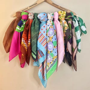 May include: Nine colourful silk scarves hanging on a wooden hanger. The scarves feature various patterns, including floral, geometric, and polka dots. The scarves are in shades of pink, green, blue, brown, and orange.