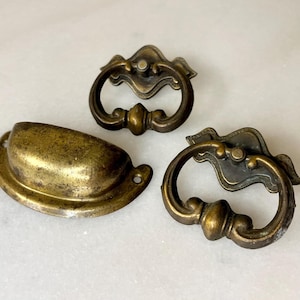 May include: Three antique brass cabinet pulls with ornate designs. Two of the pulls are ring-shaped with a decorative centre piece. The third pull is a simple, rounded, oval shape.
