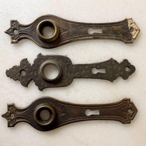 Peut inclure: Trois plaques de porte en laiton antique avec des motifs ornés. Chaque plaque a un trou de serrure et un trou rond pour un bouton.
