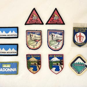 May include: A selection of vintage ski patches featuring diverse designs and text. Patches include "Killington" triangles, "Madonna Mountain" rectangles, "Banff" shields, "Mt. Snow" shields, "Garibaldi's Whistler Mountain" square, and "Nacona Ski Club" patch.