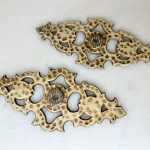 May include: Two ornate gold-toned metal decorative pieces with intricate cut-out designs. Each piece has a small round hole in the centre.