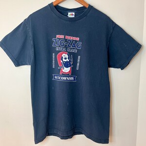 May include: A dark blue t-shirt with a graphic of a man's face with a cigarette in his mouth. The text on the shirt reads "Free Burning Zig-Zag Extra Giant Kutcorners".