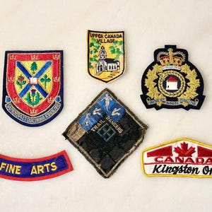 May include: A selection of embroidered patches featuring diverse designs. Includes a shield with a cross and floral motifs, a yellow patch depicting a church, a patch with a crown and wreath, a diamond-shaped patch with figures, a "FINE ARTS" patch, and a "CANADA Kingston Ont." patch.