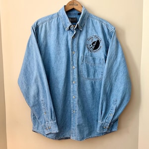 May include: Light blue denim button-down shirt with long sleeves. Features a chest pocket and a black and white embroidered patch that reads "Camp Ponache Climbing Adventures". The shirt is hanging on a wooden hanger.