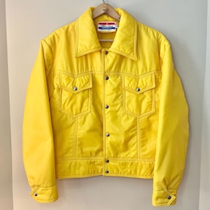 Peut inclure: Une veste matelassée jaune vif avec une fermeture à pression et deux poches à rabat. La veste a une étiquette blanche avec le texte "White Stag" sur le devant.