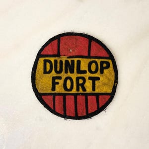 May include: A circular patch featuring the text "DUNLOP FORT" in black on a yellow panel. The patch has a red border with black lines, forming a grid design. The patch is likely vintage.