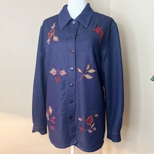May include: A dark blue denim shirt with embroidered floral and leaf designs. The shirt has a button-up front and long sleeves.