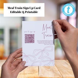 May include: A white Meal Train sign-up card with the text "Meal Train Sign Up Card Editable & Printable" at the top. The card features a QR code and the words "Meal Train" in a decorative font. The card is held by two hands.