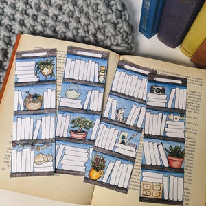 May include: Watercolor bookmarks featuring bookshelf designs with various items like plants, a cat, and a camera. The bookmarks are placed on an open book with a blue pen. A gray knitted blanket and a stack of books are in the background.