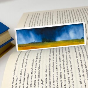 K&ouml;nnte beinhalten: Ein rechteckiger Aquarell-Lesezeichen mit einer Landschaftsszene mit dunkelblauem Himmel, gelbem Feld und gr&uuml;nen B&auml;umen. Das Lesezeichen liegt auf den Seiten eines offenen Buches, ein blaues Buch ist sichtbar.