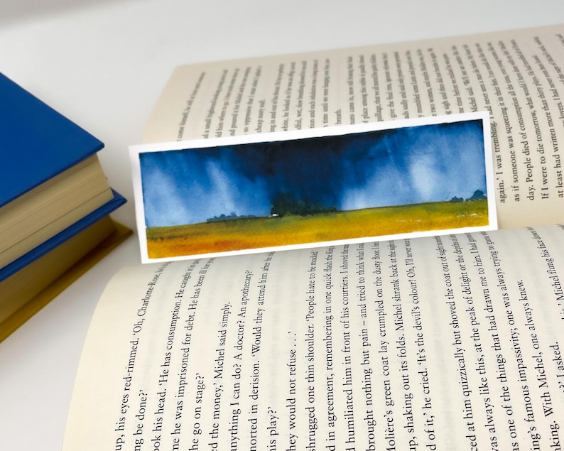 K&ouml;nnte beinhalten: Ein rechteckiger Aquarell-Lesezeichen mit einer Landschaftsszene mit dunkelblauem Himmel, gelbem Feld und gr&uuml;nen B&auml;umen. Das Lesezeichen liegt auf den Seiten eines offenen Buches, ein blaues Buch ist sichtbar.