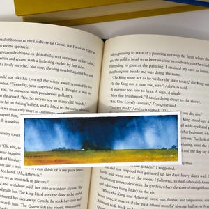 K&ouml;nnte beinhalten: Ein rechteckiger Aquarell-Lesezeichen mit einer Landschaft mit gelbem Feld, B&auml;umen und blauem Himmel mit dunklen Wolken. Das Lesezeichen liegt auf einem offenen Buch.