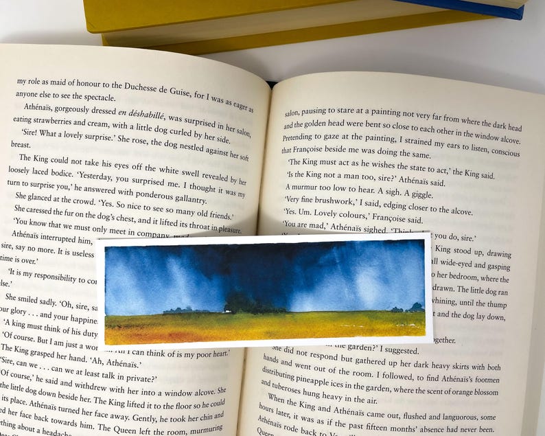 K&ouml;nnte beinhalten: Ein rechteckiger Aquarell-Lesezeichen mit einer Landschaft mit gelbem Feld, B&auml;umen und blauem Himmel mit dunklen Wolken. Das Lesezeichen liegt auf einem offenen Buch.