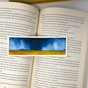 K&ouml;nnte beinhalten: Ein kleines Aquarell, das eine Landschaft mit einem gelben Feld, gr&uuml;nen B&auml;umen und einem dunkelblauen Himmel zeigt. Das Kunstwerk befindet sich vor einem offenen Buch.