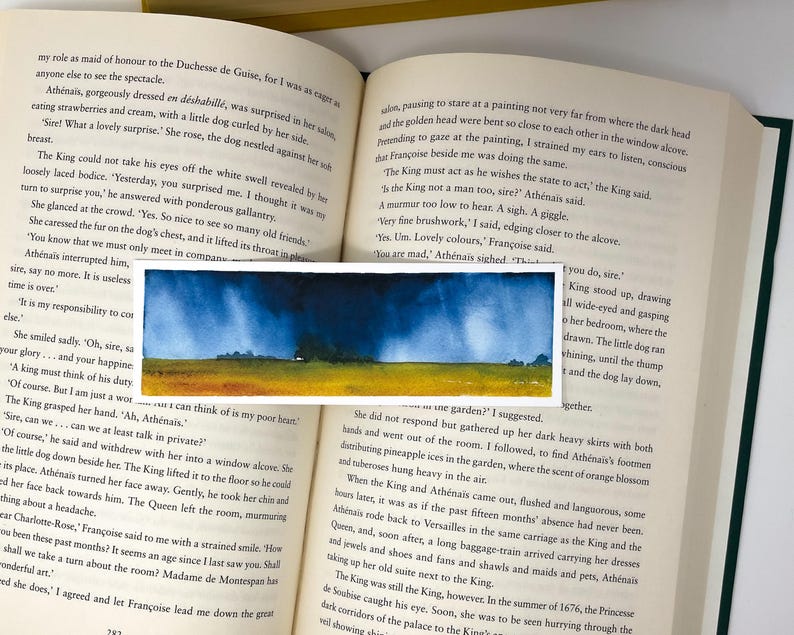 K&ouml;nnte beinhalten: Ein kleines Aquarell, das eine Landschaft mit einem gelben Feld, gr&uuml;nen B&auml;umen und einem dunkelblauen Himmel zeigt. Das Kunstwerk befindet sich vor einem offenen Buch.