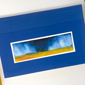 K&ouml;nnte beinhalten: Ein blaues Buch mit einem Aquarell-Landschaftsbild. Das Gem&auml;lde zeigt ein gelbes Feld, einen dunkelblauen Himmel und Regen. Weitere B&uuml;cher sind zu sehen.