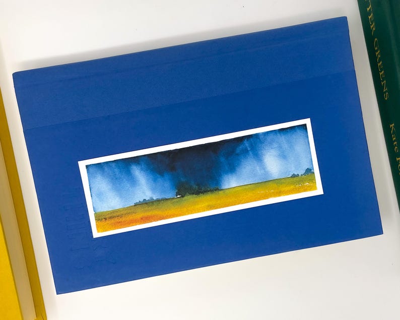 K&ouml;nnte beinhalten: Ein blaues Buch mit einem Aquarell-Landschaftsbild. Das Gem&auml;lde zeigt ein gelbes Feld, einen dunkelblauen Himmel und Regen. Weitere B&uuml;cher sind zu sehen.