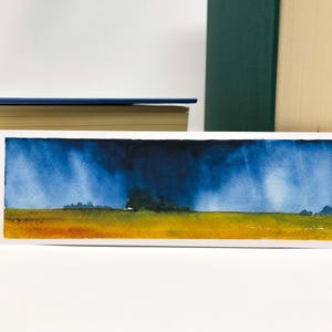 K&ouml;nnte beinhalten: Rechteckiger Aquarell-Lesezeichen mit einer Landschaftsszene. Das Kunstwerk zeigt ein gelbes und orangefarbenes Feld unter einem dramatischen blauen Himmel mit dunklen Gewitterwolken. Das Lesezeichen ist wei&szlig; umrandet.