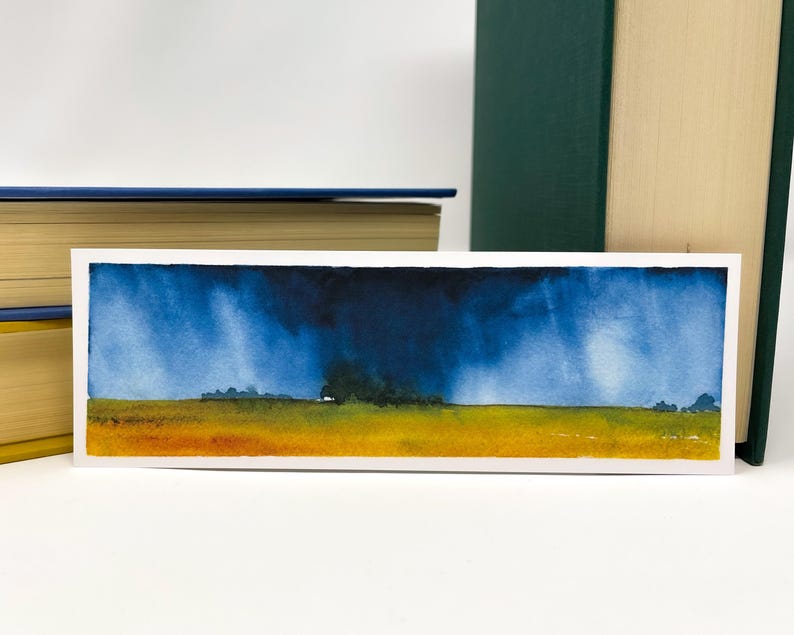 K&ouml;nnte beinhalten: Rechteckiger Aquarell-Lesezeichen mit einer Landschaftsszene. Das Kunstwerk zeigt ein gelbes und orangefarbenes Feld unter einem dramatischen blauen Himmel mit dunklen Gewitterwolken. Das Lesezeichen ist wei&szlig; umrandet.
