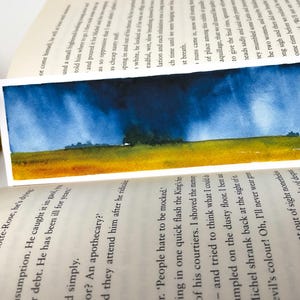 K&ouml;nnte beinhalten: Aquarell-Lesezeichen mit einer Landschaftsszene. Der obere Teil zeigt einen dunkelblauen Himmel, der in ein gelbes und gr&uuml;nes Feld &uuml;bergeht. Das Lesezeichen befindet sich in den Seiten eines Buches.