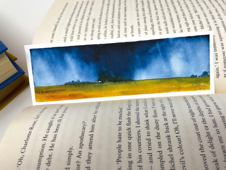 K&ouml;nnte beinhalten: Aquarell-Lesezeichen mit einer Landschaftsszene. Der obere Teil zeigt einen dunkelblauen Himmel, der in ein gelbes und gr&uuml;nes Feld &uuml;bergeht. Das Lesezeichen befindet sich in den Seiten eines Buches.