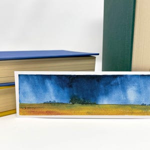 K&ouml;nnte beinhalten: Aquarell-Lesezeichen mit einer Landschaftsszene mit st&uuml;rmischem Himmel und Feld. Das Kunstwerk hat eine blaue und gelbe Farbpalette. Das Lesezeichen ist rechteckig und befindet sich neben zwei B&uuml;chern.