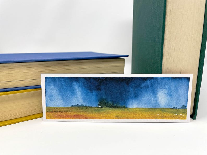 K&ouml;nnte beinhalten: Aquarell-Lesezeichen mit einer Landschaftsszene mit st&uuml;rmischem Himmel und Feld. Das Kunstwerk hat eine blaue und gelbe Farbpalette. Das Lesezeichen ist rechteckig und befindet sich neben zwei B&uuml;chern.