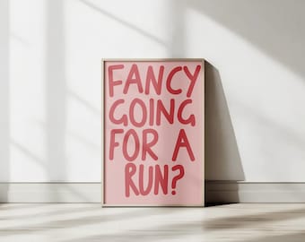 Pode incluir: Uma peça de arte de parede retangular emoldurada com um fundo rosa claro e o texto FANCY GOING FOR A RUN? escrito em uma fonte em negrito, vermelho escuro e estilo manuscrito. A moldura é de madeira clara e a impressão está centralizada.