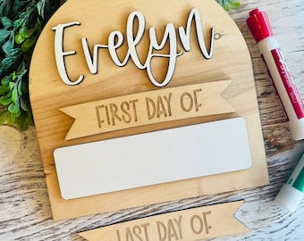 May include: A wooden sign with the name "Evelyn" written in cursive. The sign has a blank space for writing in the dates for the first and last day of school. The sign is decorated with two banners that say "First Day Of" and "Last Day Of".