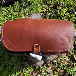 May include: A brown leather messenger bag with a flap closure, featuring a crescent moon and star emblem embossed on the front. The bag includes a small leather strap with a toggle fastener and the text GERUDO GEAR stamped on the side.