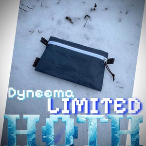 May include: A dark gray Dyneema pouch with a white zipper and black pull tab, lying on snow. The text "Dyneema LIMITED HOTT" is displayed in pixelated blue and white font.