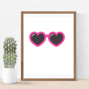 May include: A framed print featuring a pair of heart-shaped sunglasses with a bright pink frame and black lenses. The artwork is set against a white background, with a cactus in a white pot to the left.
