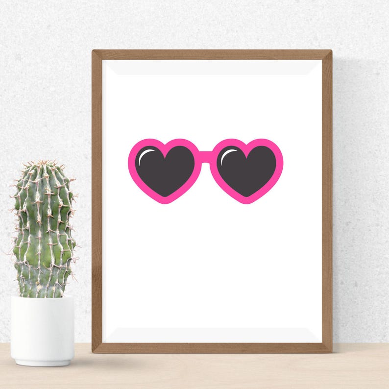 May include: A framed print featuring a pair of heart-shaped sunglasses with a bright pink frame and black lenses. The artwork is set against a white background, with a cactus in a white pot to the left.