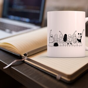 May include: White ceramic mug with a black line drawing of seven women standing back to back with their arms around each other. The women are wearing skirts and have their hair pulled back.